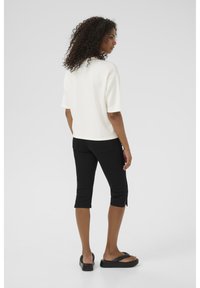 White short-sleeve top, black cropped pants with side zippers, and black sandals. Model facing back, hair in loose curls, neutral background.