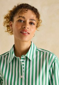 Grünes und weiß gestreiftes Hemd mit Knöpfen und Kragen. Der Stoff ist glatt und weist gleichmäßige vertikale Streifen auf. Enthält Knopfdetails am Halsausschnitt.