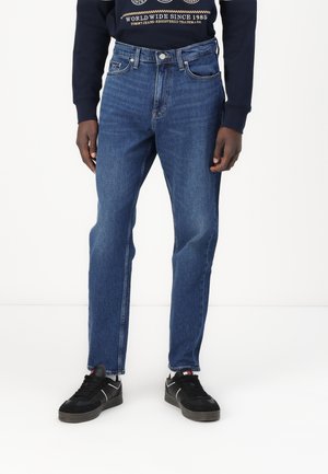 Hombre con vaqueros azul medio de pierna recta, zapatillas negras y sudadera azul marino con texto gráfico blanco y dorado.