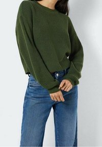 Pull en maille vert foncé avec un col rond et des manches longues légèrement oversize ; associé à un jean bleu à jambes droites.