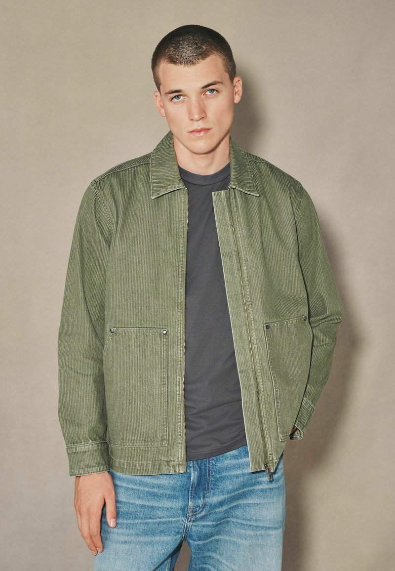 Olívazöld kordbársony dzseki cipzáros elülső résszel, két oldalsó zsebbel és galléros nyakkivágással, fekete póló és kék farmer felett viselve.