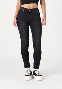 Jeans negros ajustados con una cintura alta, que cuentan con un cierre de botón en la parte frontal, un diseño clásico de cinco bolsillos y un dobladillo ligeramente deshilachado. Combinados con zapatillas negras y blancas.