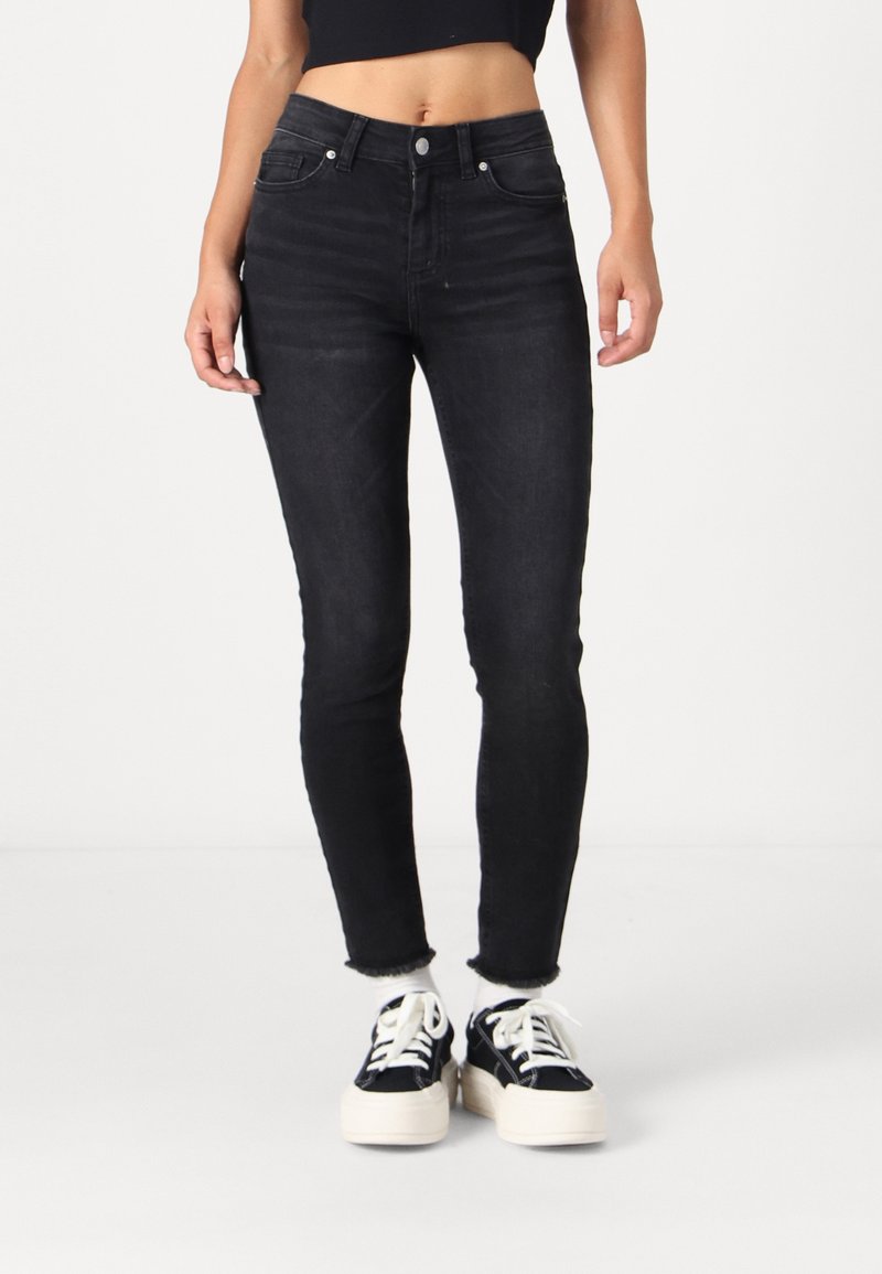 Jeans negros ajustados con una cintura alta, que cuentan con un cierre de botón en la parte frontal, un diseño clásico de cinco bolsillos y un dobladillo ligeramente deshilachado. Combinados con zapatillas negras y blancas.