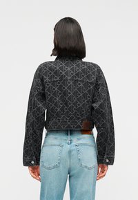 Κοντό μαύρο τζιν μπουφάν με επαναλαμβανόμενο σχέδιο, με κούμπωμα με κουμπιά και δερμάτινη ετικέτα στην πλάτη. Φορεμένο με ανοιχτόχρωμο μπλε τζιν.