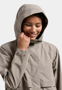 Frau lächelt, während sie die Kapuze einer hellbeigen wasserdichten Jacke mit knöpfbaren Manschetten vor einem einfarbigen Hintergrund anpasst.