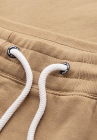 Vita della tuta beige con texture a coste e cordino bianco infilato attraverso occhielli in metallo nero con testo.