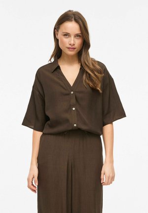 Femme aux longs cheveux portant une chemise marron ample à boutons et un pantalon large assorti, debout avec une posture détendue devant un fond blanc.