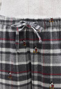 Pantalon de pyjama à carreaux gris et blanc avec des lignes rouges et de petits motifs de Mickey Mouse, attaché à la taille avec un nœud en tissu.