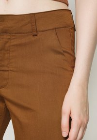 Pantaloni in misto cotone marroni con una texture liscia, caratterizzati da una parte frontale piatta, tasche laterali e passanti per la cintura, mostrati con una mano appoggiata in vita.
