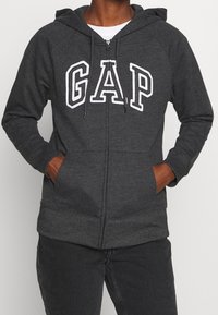 Sudadera gris con cremallera y logo blanco "GAP" en el pecho, capucha con cordón, dos bolsillos frontales, y puños y dobladillo acanalados. Textura de tela suave.