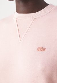 Personne à la peau foncée portant un sweat-shirt à col rond rose clair avec un petit logo crocodile rose brodé sur la poitrine.