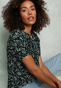 Blusa negra con estampado floral que presenta mangas cortas, un escote con volantes y un patrón de pequeñas flores azules y blancas sobre un fondo oscuro.
