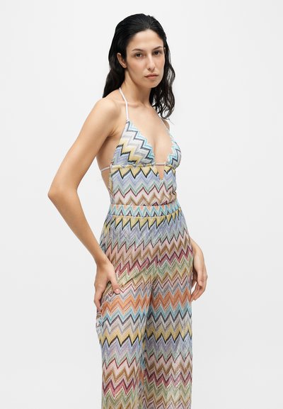 Femme aux longs cheveux foncés porte une combinaison sans manches à motif zigzag multicolore avec fines bretelles halter, posant avec une main sur la hanche.