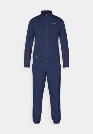 Mørkeblå joggedress laget av lett materiale. Inkluderer en jakke med glidelås og høy krage, samt matchende bukser med elastisk midje.