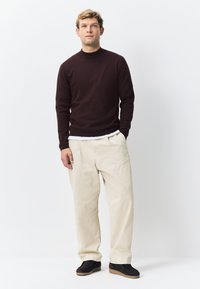 Burgunderfarvet strikket trøje med lange ærmer, cremeløse bukser med vide ben og sorte sko med tekstureret sål; modellen står foran en ensartet hvid baggrund.