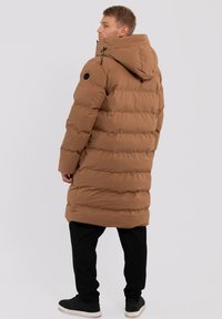 Braune Pufferjacke mit Kapuze, ausgestattet mit einem Steppmuster, verstellbaren Kordelzügen und einem schwarzen Logo-Patch auf dem oberen Arm, getragen mit schwarzen Hosen.