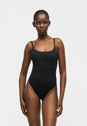 Kobieta z plecionymi włosami, ubrana w czarny bezrękawnik body, stojąca na tle jednolitego jasnoszarego tła, zwrócona przodem.