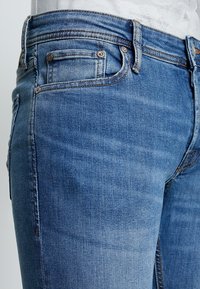 Jeans de denim azul claro con un corte ajustado, que cuentan con un diseño de cinco bolsillos, herrajes de cobre y un ligero desgaste en la tela.