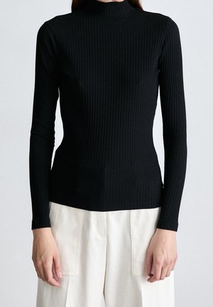 Frau trägt einen figurbetonten schwarzen, gerippten Rollkragenpullover mit langen Ärmeln und elfenbeinfarbene, hoch taillierte Hosen vor neutralem Hintergrund.