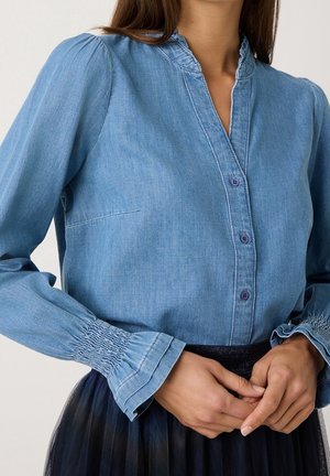 Woman wearing light blue denim blouse with ruffled cuffs and dark pleated skirt, hands clasped at waist.