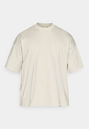 Svetlo bež kratka majica s kratkimi rokavi iz bombaža, z okroglim izrezom in rahlo preveliko zasnovo ter črtastim poudarkom na strani.