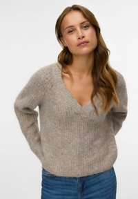 Pull en maille beige clair avec un col en V, manches longues et une finition texturée. Associé à un jean en denim bleu.