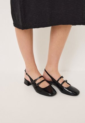 Nogi w czarnych lakierowanych butach typu slingback z dwoma paskami na klamry i niskimi obcasami typu blok, połączone z czarną dzianą spódnicą.