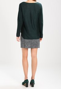 Donkergroen gebreide trui met geribbelde textuur, gecombineerd met een zwart-wit houndstooth rok, gedragen met groene hoge hakken.