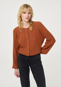 Geribbelde, cropped cardigan in gebrandoranje met gouden knopen, ronde halslijn en lange mouwen. Gecombineerd met zwarte high-waisted jeans.