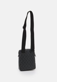 Černá ramenní taška vyrobená z potištěné látky s opakujícím se logem. Vybavena nastavitelným černým popruhem a zipovým uzávěrem.