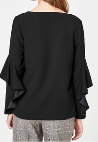 Black top featuring flared sleeves and a square neckline. The fabric appears smooth. Worn with patterned, light-check pants.