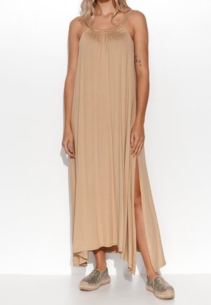 Mujer con vestido maxi sin mangas beige con abertura lateral y alpargatas metálicas sin cordones, de pie contra un fondo blanco.