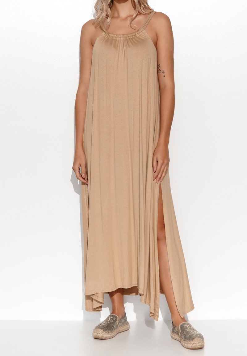 Femme portant une robe maxi beige sans manches avec une fente latérale et des espadrilles métalliques à enfiler, debout devant un fond blanc.
