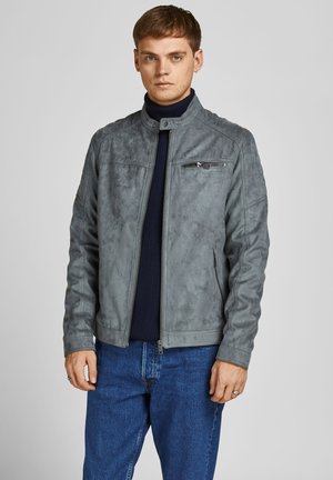 Chaqueta de ante sintético gris con cuello alzado, bolsillos con cremallera y superficie texturizada, llevada sobre un suéter azul oscuro y jeans.