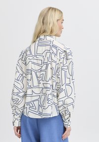 Camicia a maniche lunghe in tessuto crema con un motivo geometrico blu, dal taglio rilassato e polsini con bottone. Collo semplice e dettagli delle cuciture delle spalle.