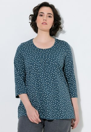 Mujer con el pelo corto y oscuro que lleva una camisa azul de manga larga con pequeños patrones de triángulos blancos, de pie sobre un fondo blanco.