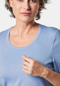 Lichtblauw T-shirt met korte mouwen en een ronde hals, gemaakt van zachte stof. Een persoon raakt voorzichtig het materiaal van het shirt aan met één hand.