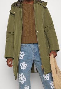 Parka acolchada de color verde oliva con capucha estampada, combinada con un suéter de punto texturizado marrón y jeans de color azul claro con diseños florales blancos.
