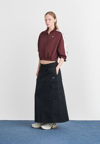 Bordo skrátená bunda s bielymi bočnými pruhmi, skombinovaná s čiernou maxi sukňou s bočnou kapsou a logom Nike. Biely tenisky dotvárajú outfit.