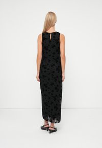 Zwarte mouwloze maxijurk met een bloemenpatroon, sleutelgatdetail op de rug, vloeiende stof en een transparante overlay. Gecombineerd met zwarte haksandalen.