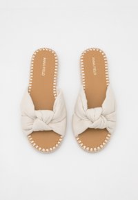 Beige stof slide sandalen met een geknoopte design aan de voorkant, een bruine zool en decoratieve stiksels rond de randen.