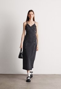 Vestido de denim preto com corpete justo, alças finas e detalhes de tachas. Comprimento até a panturrilha com uma silhueta reta, acompanhado de uma bolsa preta.