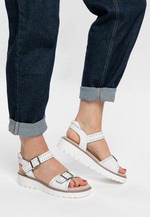 Weiße Plateau-Sandalen mit verstellbaren Riemen, Nietenakzenten und einer strukturierten Sohle. Getragen zu hochgekrempelten dunklen Jeans.