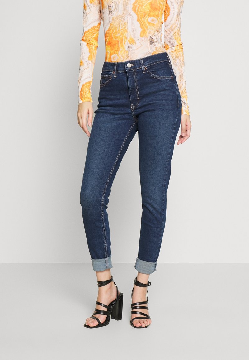 Jeans de mezclilla de tiro alto y color azul oscuro, con corte ajustado y dobladillos enrollados. Presentan costuras doradas y un cierre frontal con botón. Combinados con tacones negros de tiras.