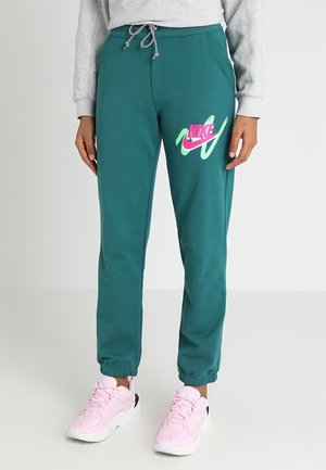 Pantalon de survêtement Nike bleu sarcelle avec logo rose et vert sur la cuisse gauche, porté avec un sweat-shirt gris clair et des chaussures de sport roses.