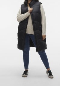 Persona che indossa un lungo gilet puffo nero sopra un maglione beige, jeans blu e sneaker bianco e nero, in piedi di fronte a uno sfondo bianco uniforme.