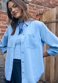 Femme debout en plein air, portant une chemise bleu clair par-dessus un haut assorti, avec un foulard bleu autour du cou et les bras pliés derrière le dos.