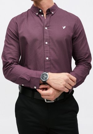 Homme portant une chemise longue prune boutonnée avec un petit logo blanc d'oiseau, un pantalon noir, une ceinture noire et une montre noire ajustant sa manche.