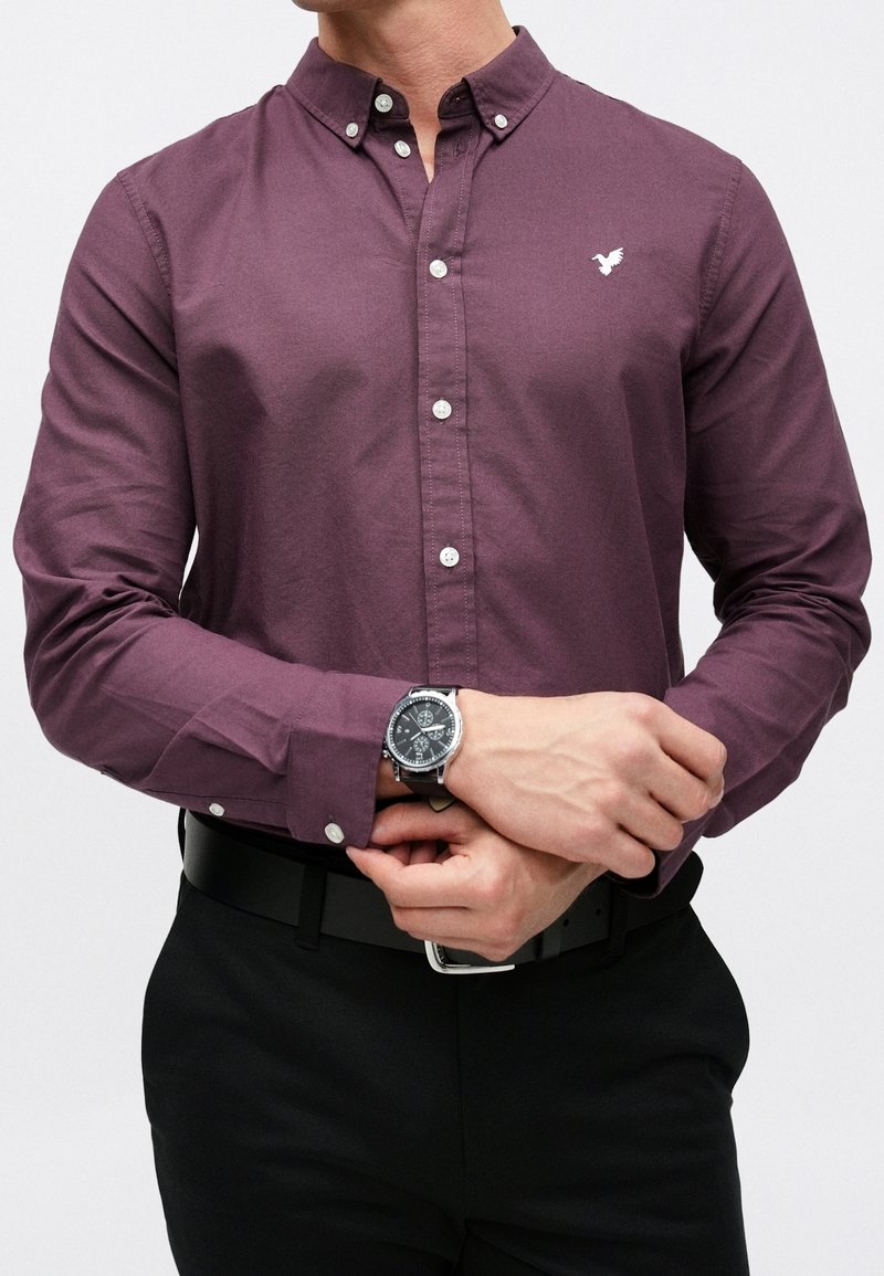Homme portant une chemise longue prune boutonnée avec un petit logo blanc d'oiseau, un pantalon noir, une ceinture noire et une montre noire ajustant sa manche.