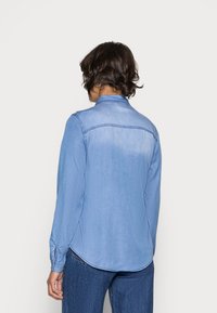 Light blue denim shirt with a curved hem, button cuffs, and a back yoke seam. Features a soft texture and casual design.